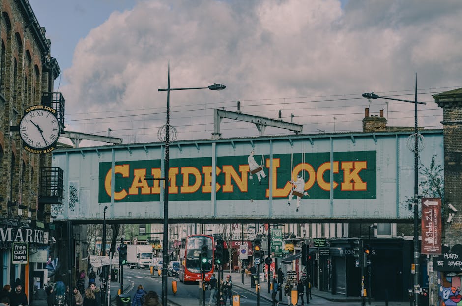 A street scene near Camden Market showing a large painted metal bridge with a prominent sign reading 'Camden Lock' in bold yellow letters on a dark green background. The bridge is suspended above a bu